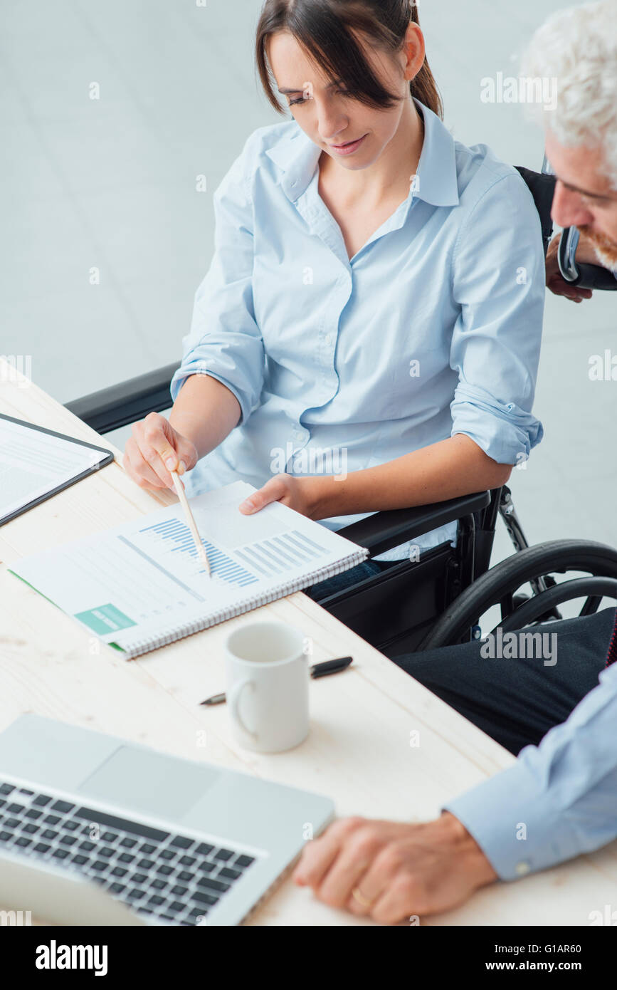 Giovane donna di affari su una sedia a rotelle a lavorare alla scrivania in ufficio e il controllo di documenti cartacei con il suo collega di sesso maschile, handicap Foto Stock
