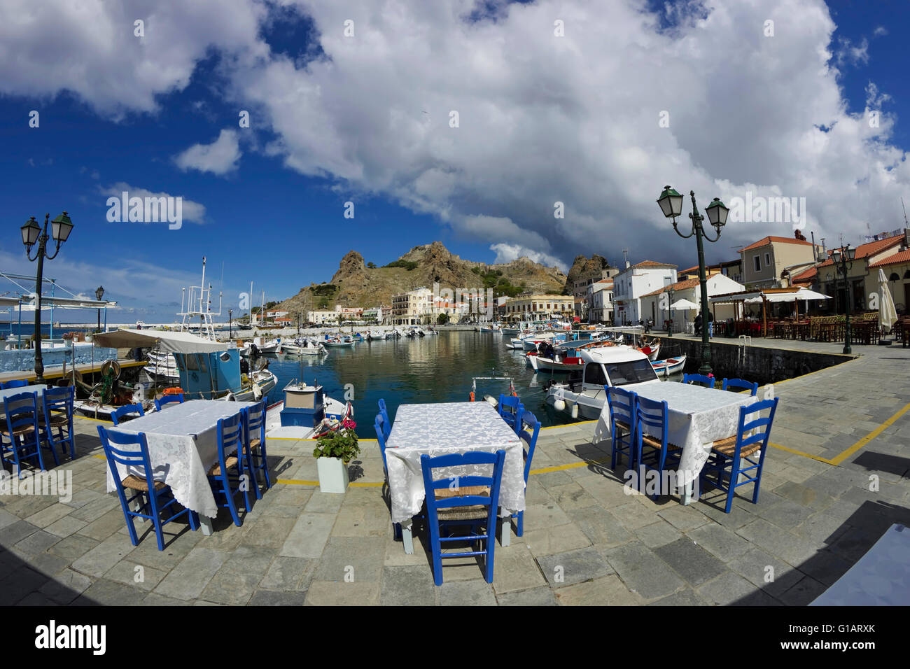 "Glaros' ristorante tavoli con sedili blu, affacciato sul castello e la città di Quay & drammatico le nuvole. Limnos, Grecia Foto Stock