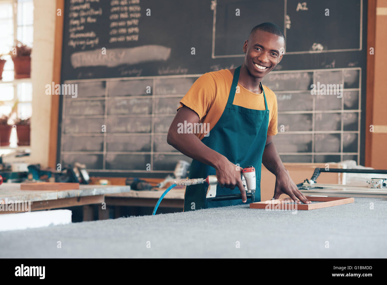 Ritratto di un bel giovane artigiano della discesa africana lavora con un utensile in una incorniciatura workshop, sorridente largamente a Foto Stock