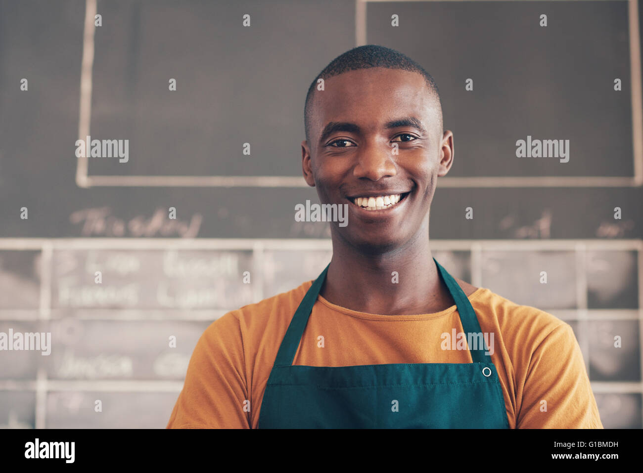 Testa e spalle il ritratto di un bel uomo africano di indossare un grembiule e sorridente ampiamente con la telecamera, mentre in piedi la Foto Stock