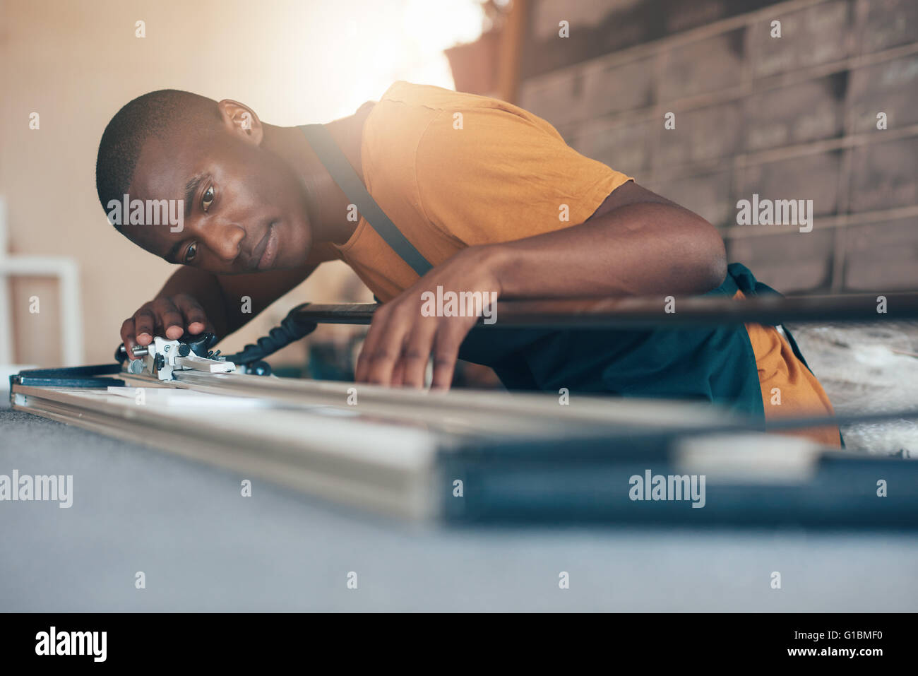 Bassa angolazione di un concentrato giovane artigiano della discesa africana, lavorando in una incorniciatura officina, utilizzando un utensile di taglio con s Foto Stock