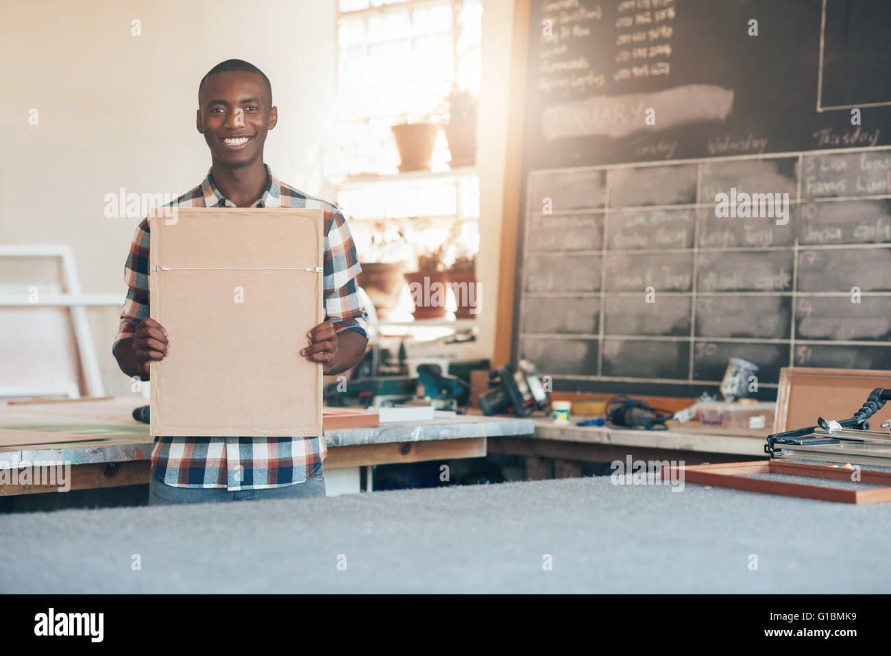 African proprietario di una piccola azienda di produzione di cornici, sorridente alla telecamera tenendo una cornice per una foto fatta a Foto Stock