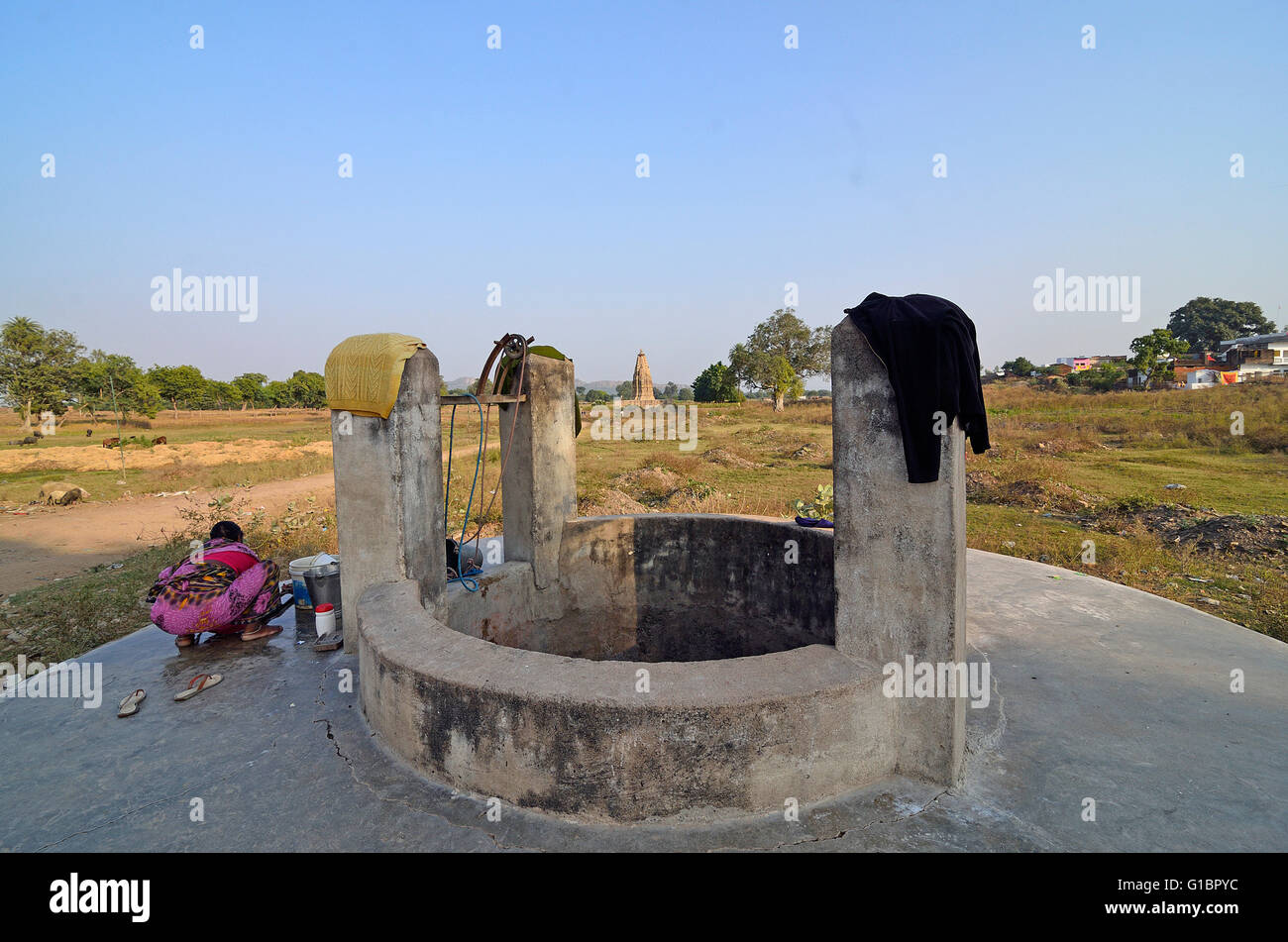 Un profondo pozzo nel villaggio di Khajuraho con una donna rurale, lavaggio con Javari temple nel lontano sfondo, Madhya Pradesh, India Foto Stock