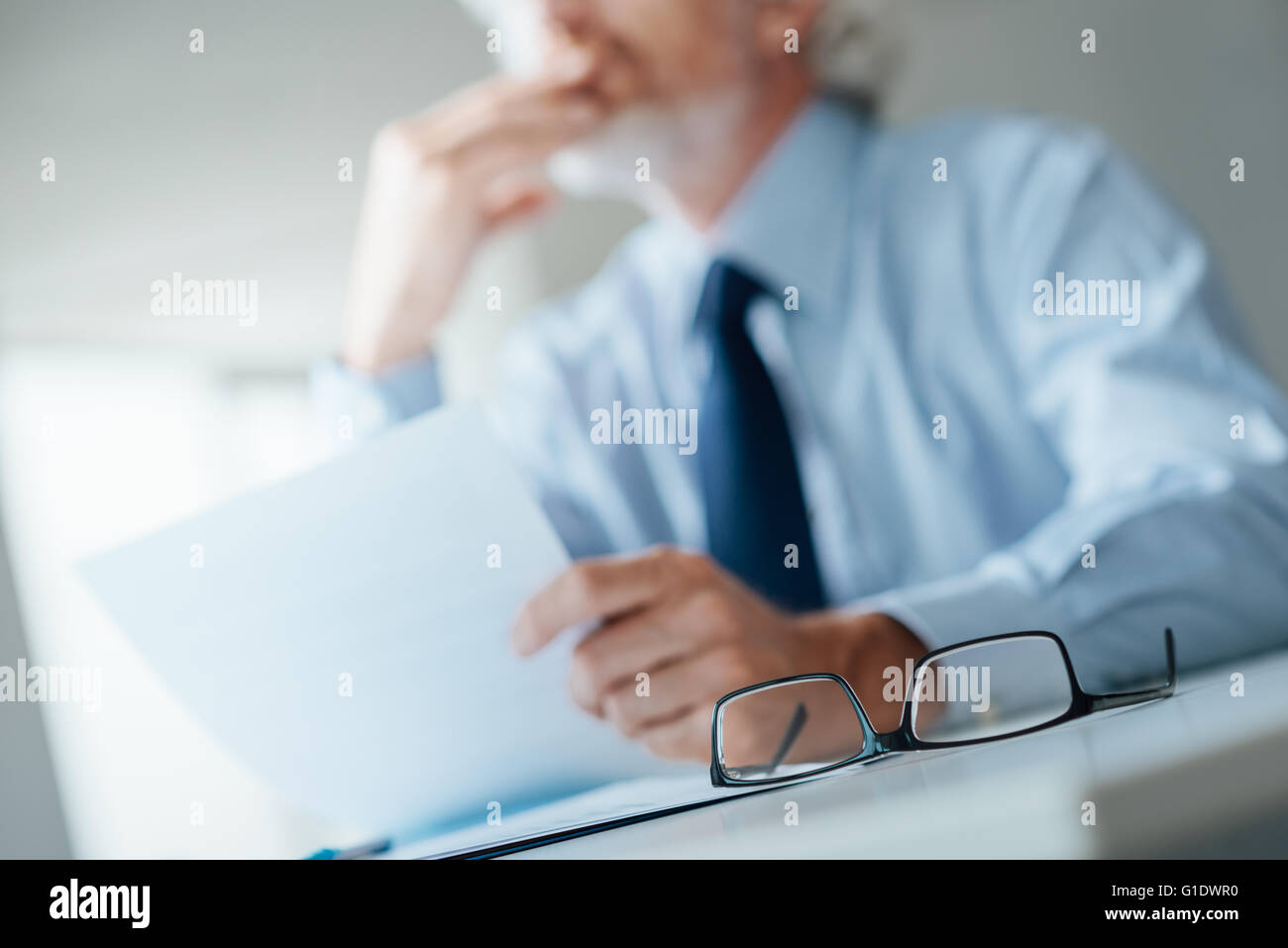 Malinconici imprenditore con mano sul mento che guarda lontano e in possesso di un documento, il fuoco selettivo bicchieri in primo piano Foto Stock