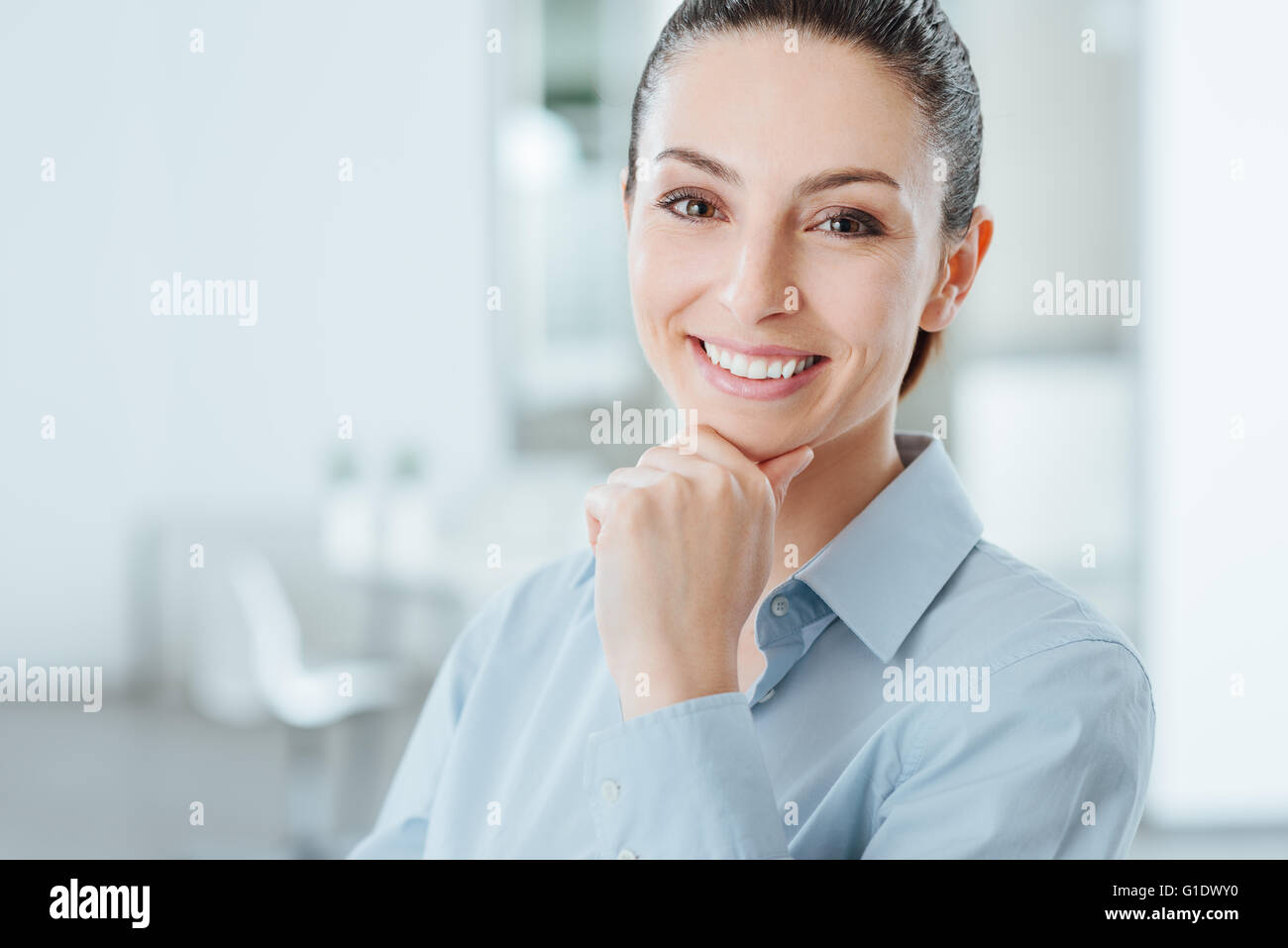Piuttosto giovane donna in posa con la mano sul mento e sorridente alla fotocamera, sala interna su sfondo Foto Stock
