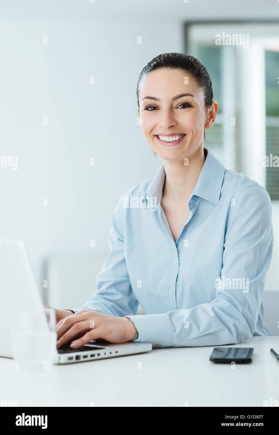 Sorridente giovane imprenditrice lavorare alla scrivania in ufficio e digitando su un portatile, è guardando la fotocamera Foto Stock