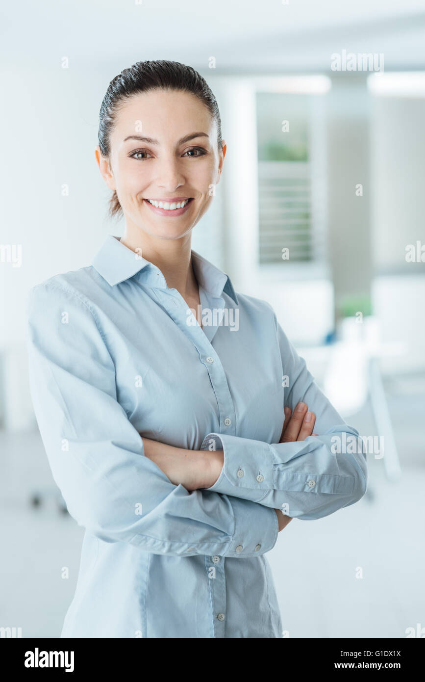 Bella fiducioso business donna posa con bracci incrociati, sorridente in telecamera e in piedi nel suo ufficio Foto Stock