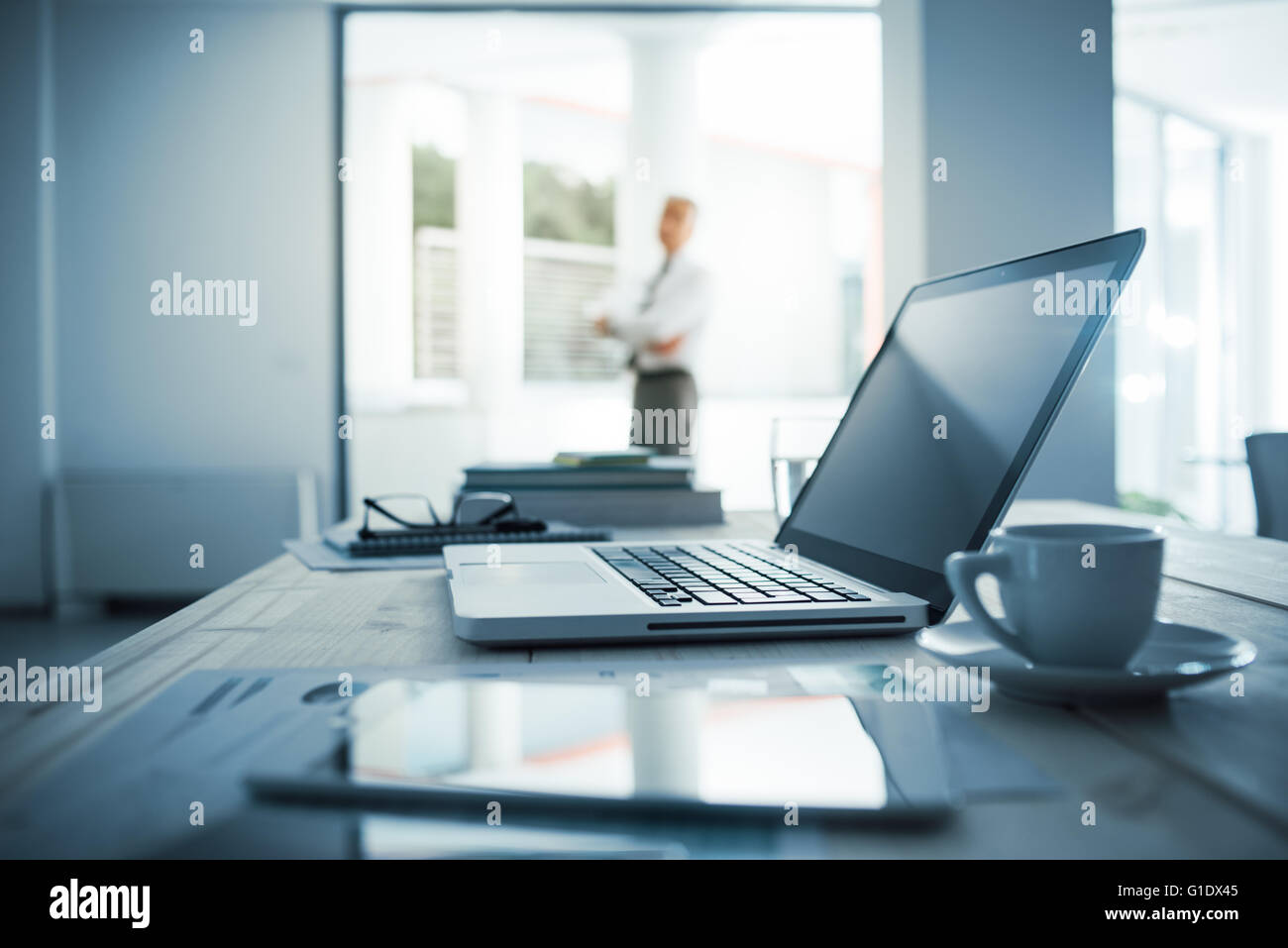 Imprenditore in piedi con le braccia incrociate nel suo ufficio, scrivania con computer portatile sul primo piano di messa a fuoco selettiva Foto Stock