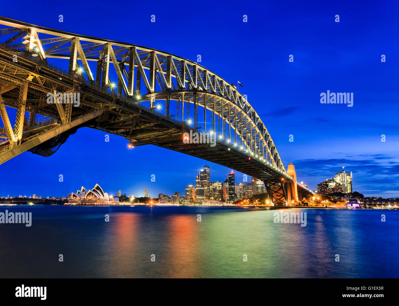 Vista laterale del Sydney Harbour Bridge verso la città CBD, rocce e Circular Quay al tramonto in tutta sfocata porto riflettente Foto Stock