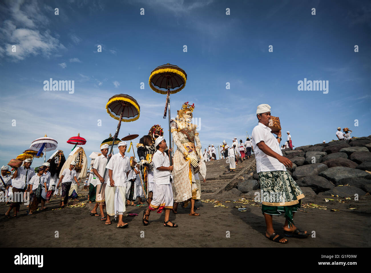 Offerte di religiosi, ombrelloni e alti personaggi mitologici nella cultura Balinese sono trasportati lungo una spiaggia di Bali Foto Stock