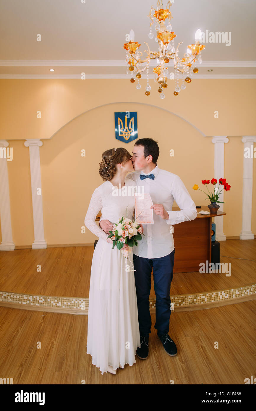 Felice coppia di sposi kissing al registro di sistema tenendo premuto il certificato di matrimonio Foto Stock
