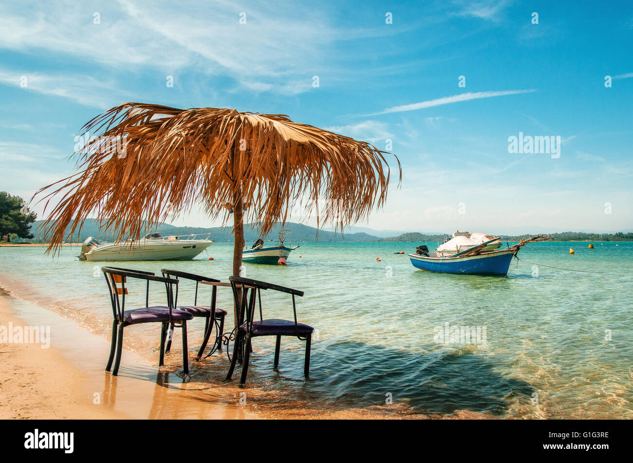 Sedie vuote sotto ombrellone in paglia su una spiaggia di sabbia vicino ad un mare pulito su uno sfondo di barche e yacht in Halkidiki, Grecia Foto Stock