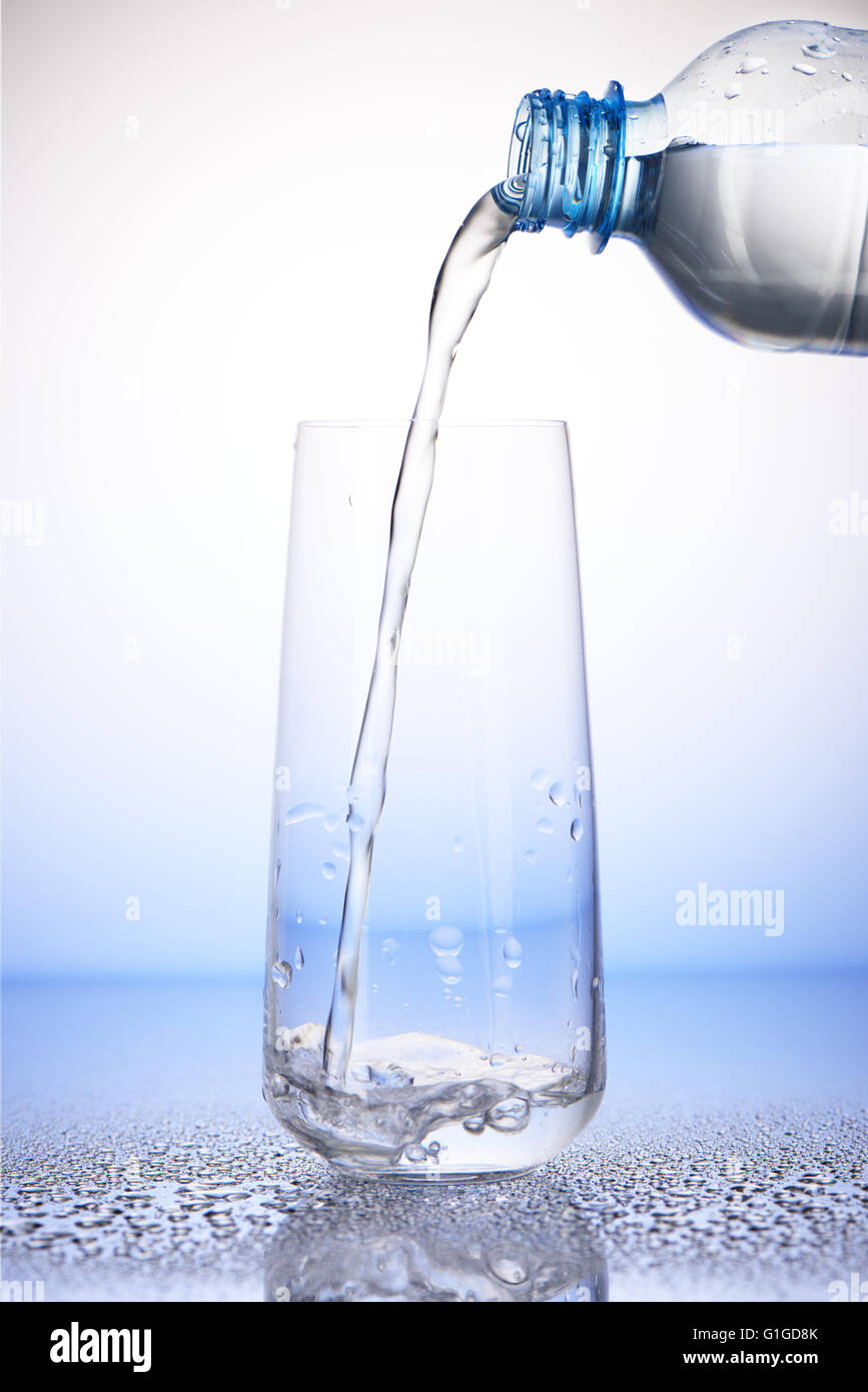 Versare acqua dalla bottiglia di plastica in un terzo bicchiere con la riflessione su gocce di acqua su bianco e blu Foto Stock