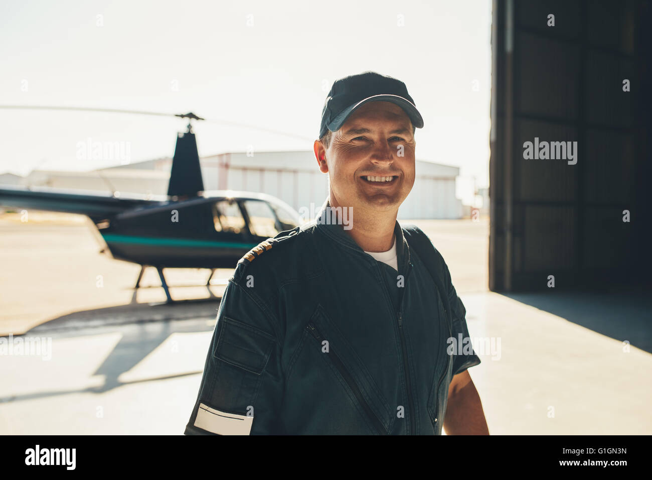 Ritratto di felice pilota maschio in piedi in hangar aereo con un elicottero in background Foto Stock