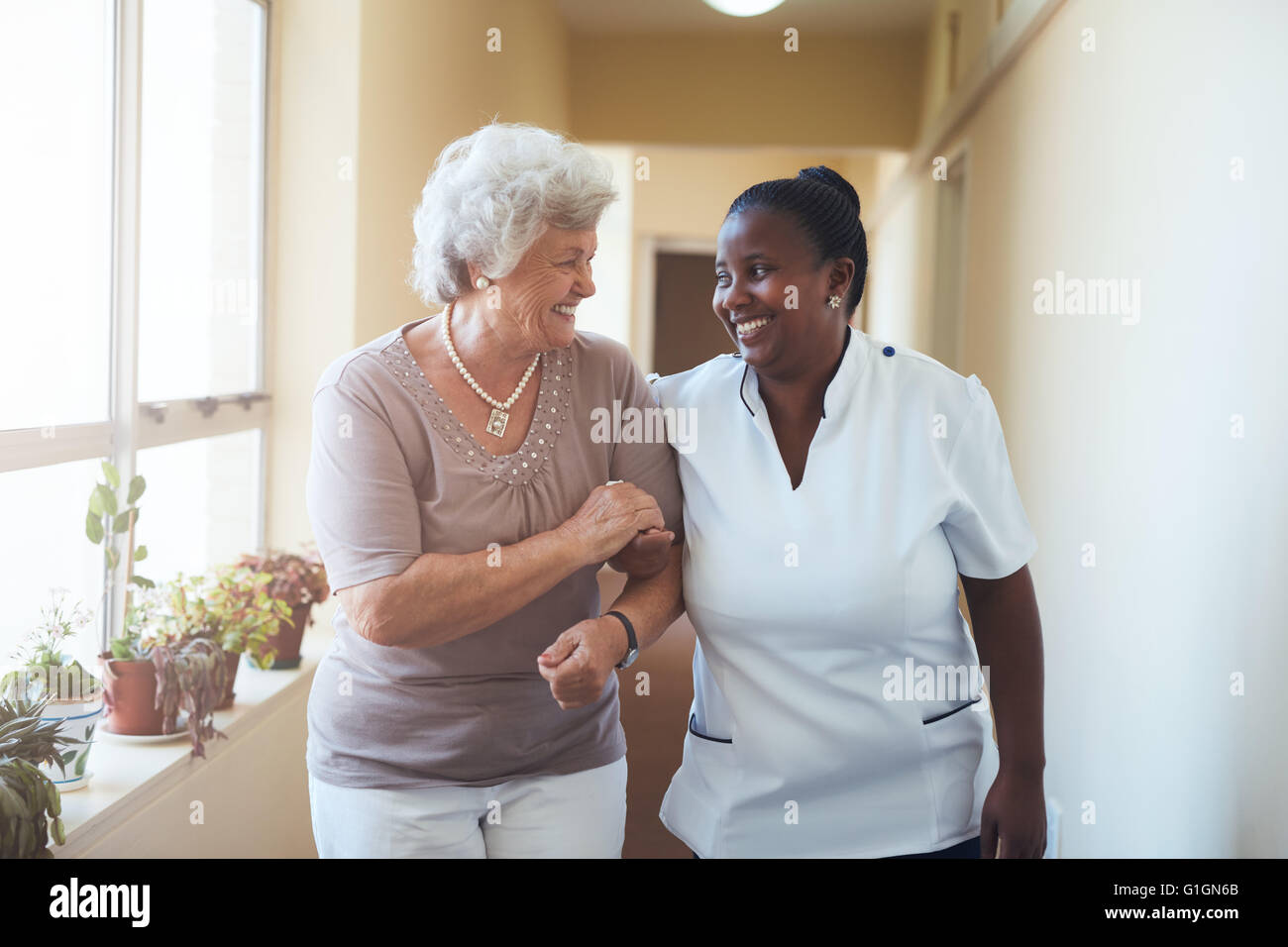 Ritratto di sorridere home caregiver e senior donna camminare insieme attraverso un corridoio. Healthcare lavoratore prendersi cura di elderl Foto Stock