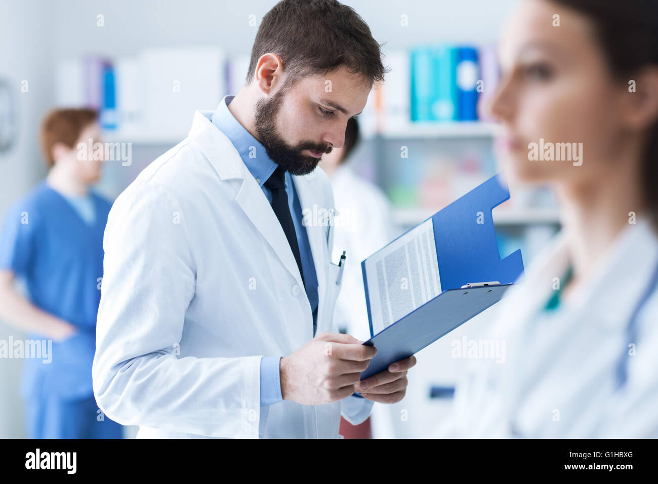 Fiducioso medico controllare i record medici su di un fermaglio nel suo ufficio, il personale medico sullo sfondo, il concetto di assistenza sanitaria Foto Stock