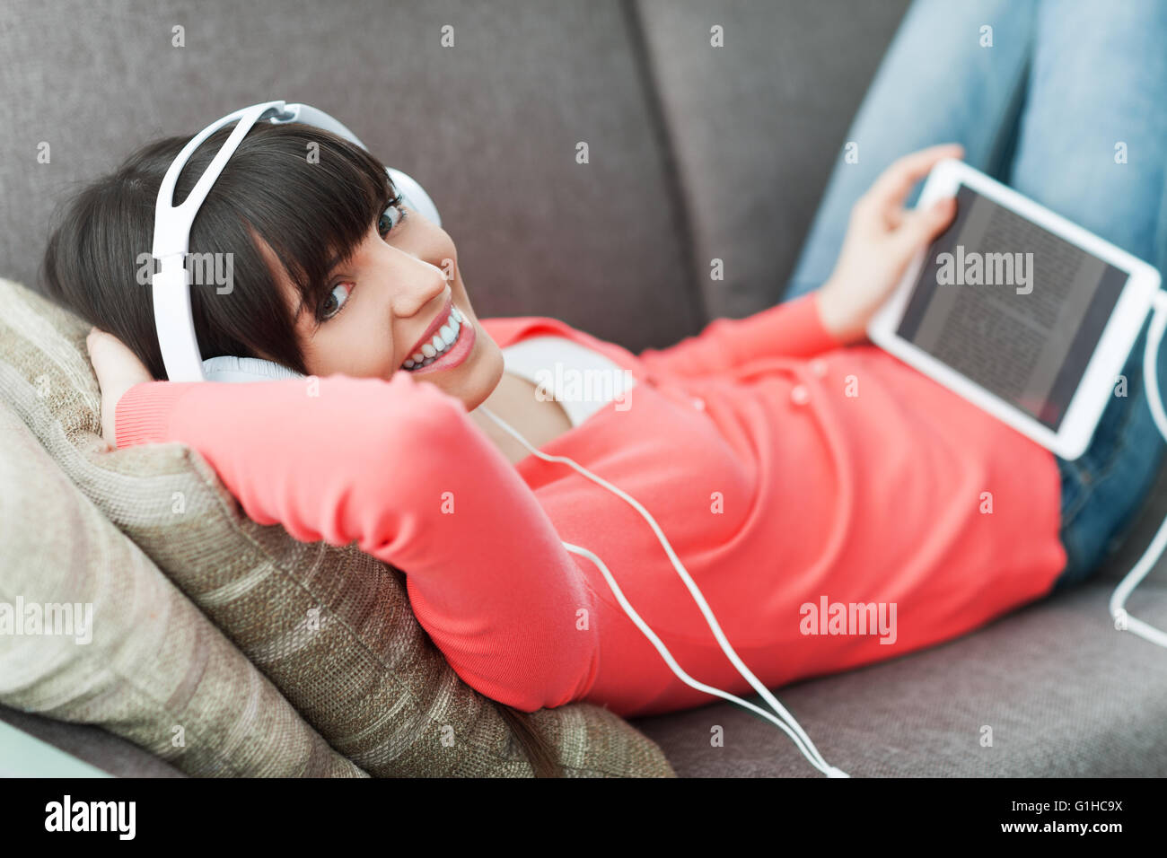Sorridente giovane donna in un momento di relax a casa sul lettino, indossa le cuffie, utilizzando una tavoletta digitale e la visione di un video online Foto Stock
