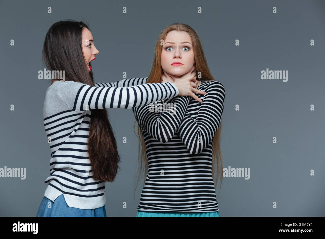 Mad arrabbiato giovane donna in piedi e soffoca scioccato giovane femmina su sfondo grigio Foto Stock