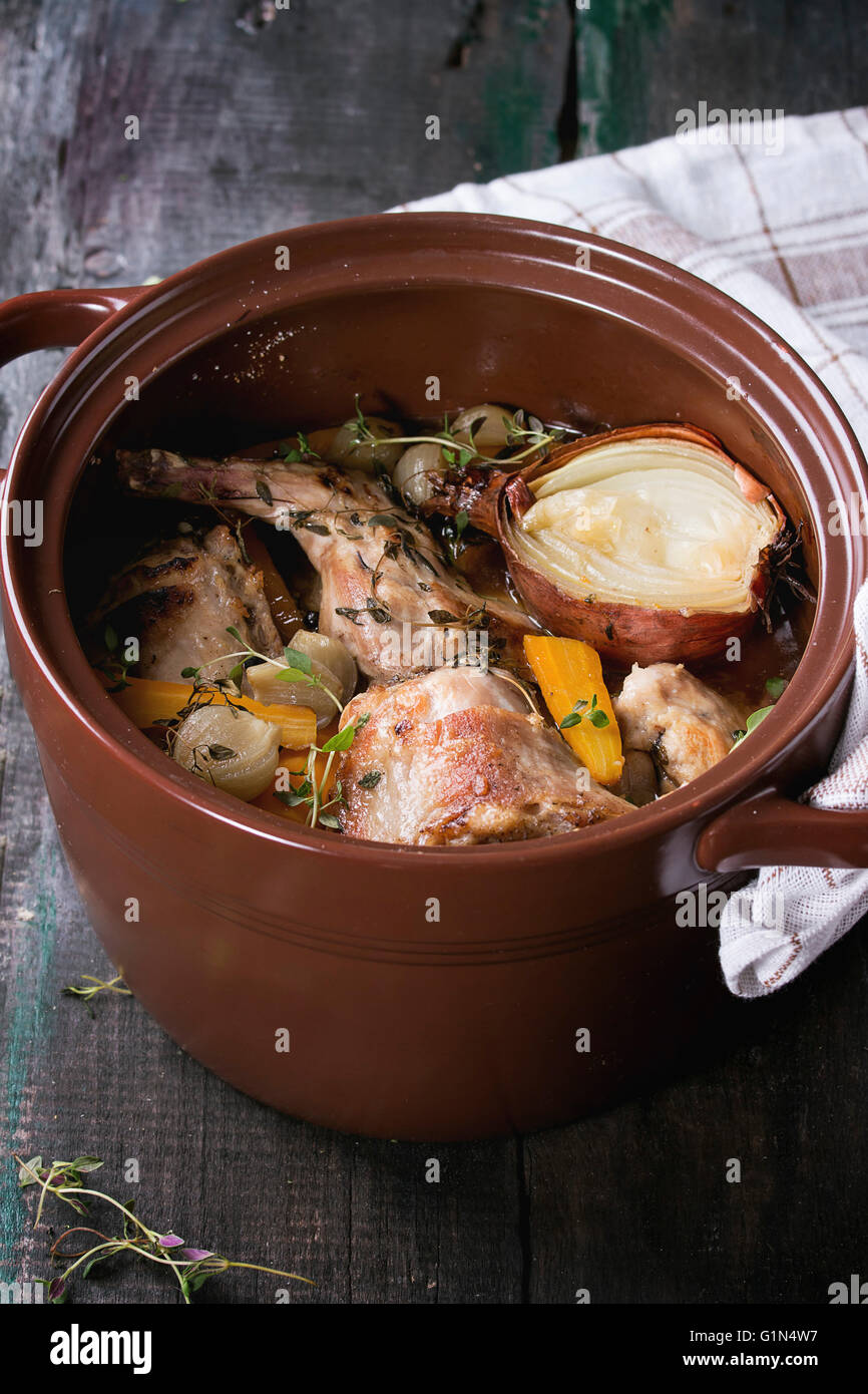 Brown vaso di ceramica con stufato di coniglio con bouillon, le verdure e le erbe aromatiche, servita con asciugamano da cucina sopra il vecchio tavolo in legno. Rusti Foto Stock