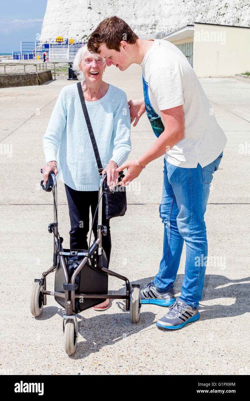 Un'anziana donna disabile utilizzando un Rollator aiuti a piedi aiutato da suo nipote, Brighton, Sussex, Regno Unito Foto Stock