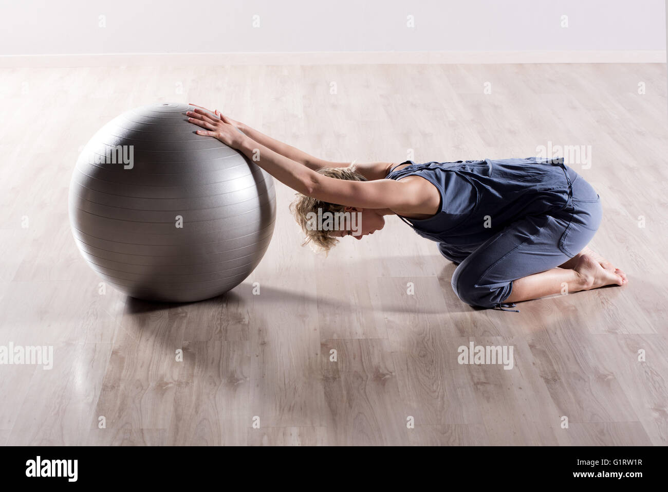 Unica donna flessibile stretching le spalle con le braccia tese sulla sommità di argento sfera fitness su pavimenti in legno studio piano Foto Stock