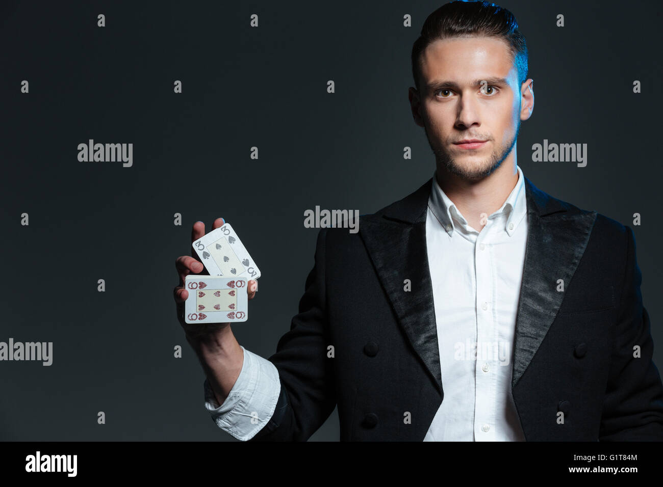 Grave giovane mago e permanente tenendo due carte da gioco su sfondo grigio Foto Stock