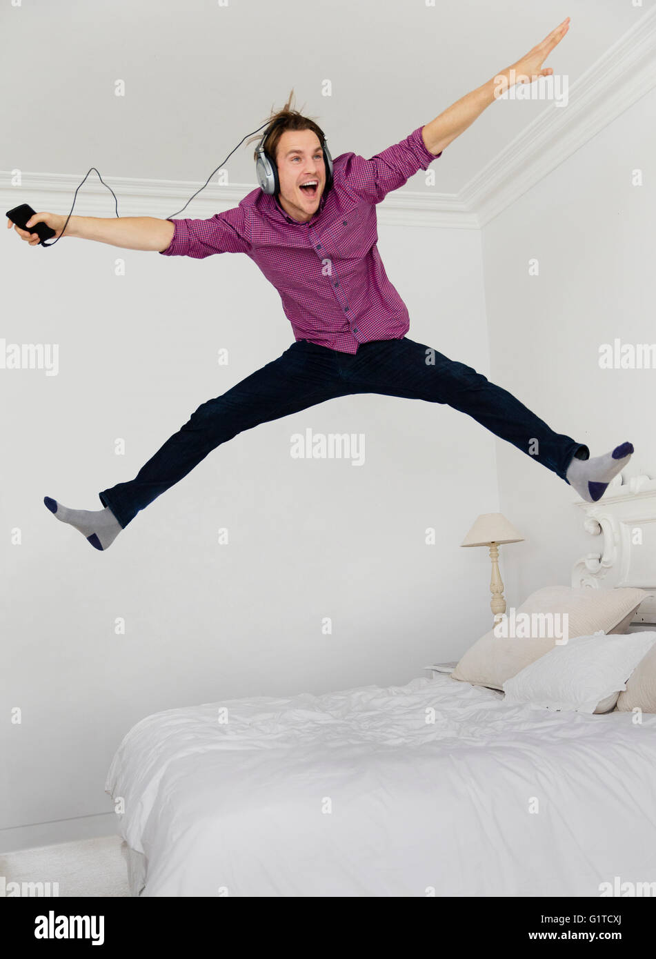 Giocoso uomo saltare sul letto con gambe divaricate per ascoltare musica con il lettore mp3 e cuffie Foto Stock