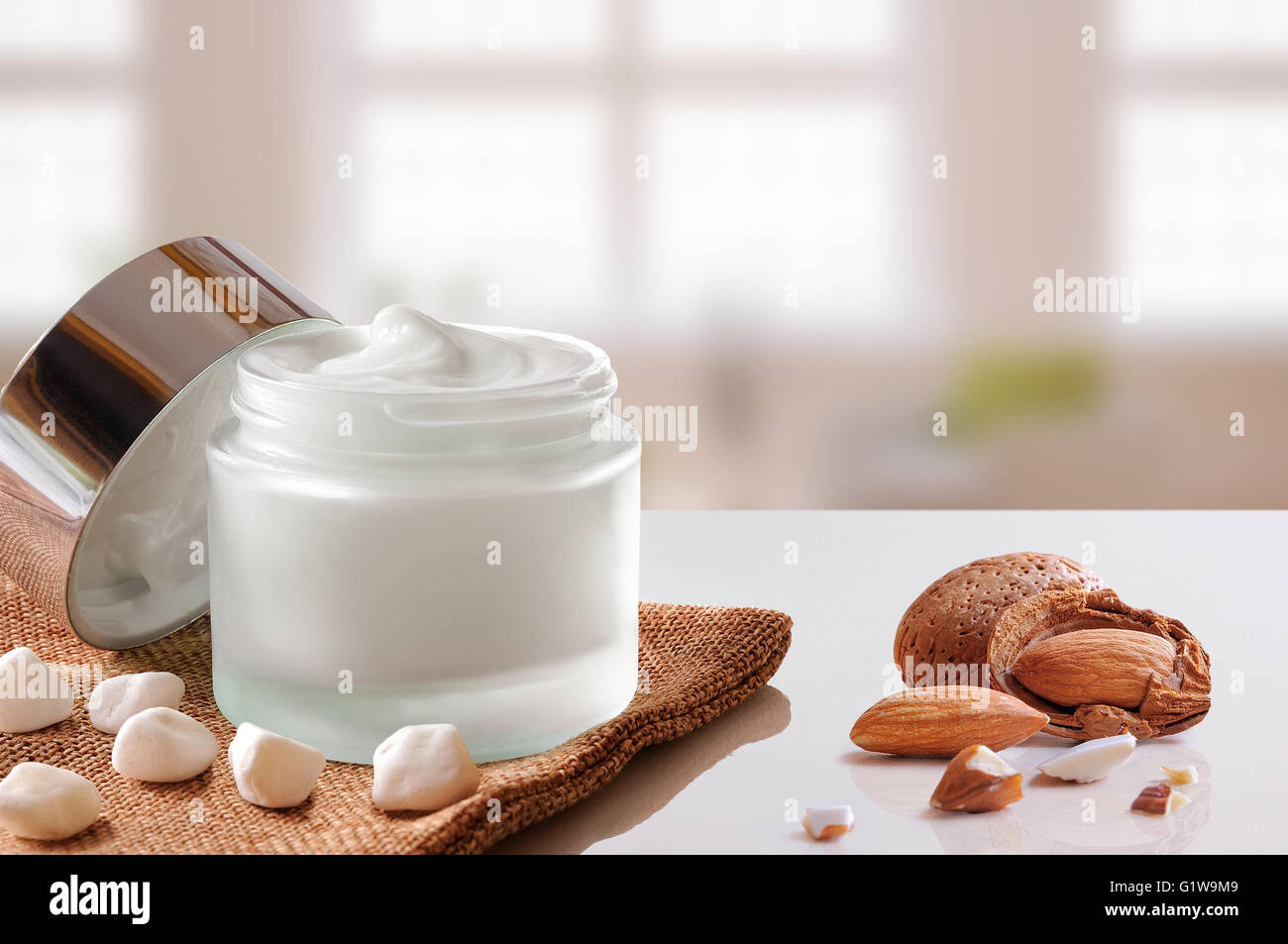 Vetro vaso aperto con viso o corpo idratante mandorle su tela. Con coperchio, con piccole pietre e mandorle. Sfondo di Windows. Foto Stock