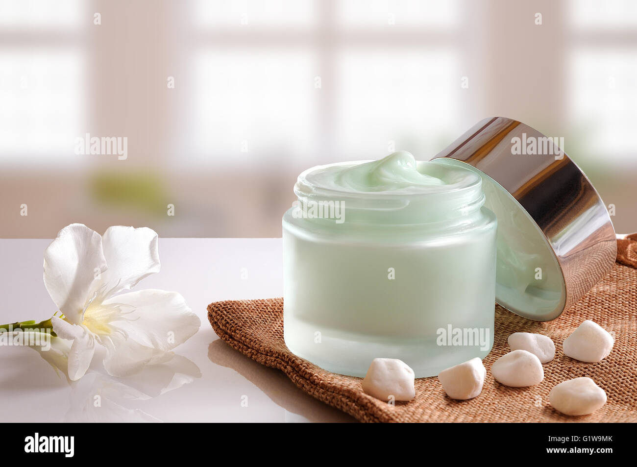 Vetro vaso aperto con viso o corpo crema sulla tela ruvida. con coperchio, pietre e fiori. Sfondo di windows. Vista frontale. Foto Stock