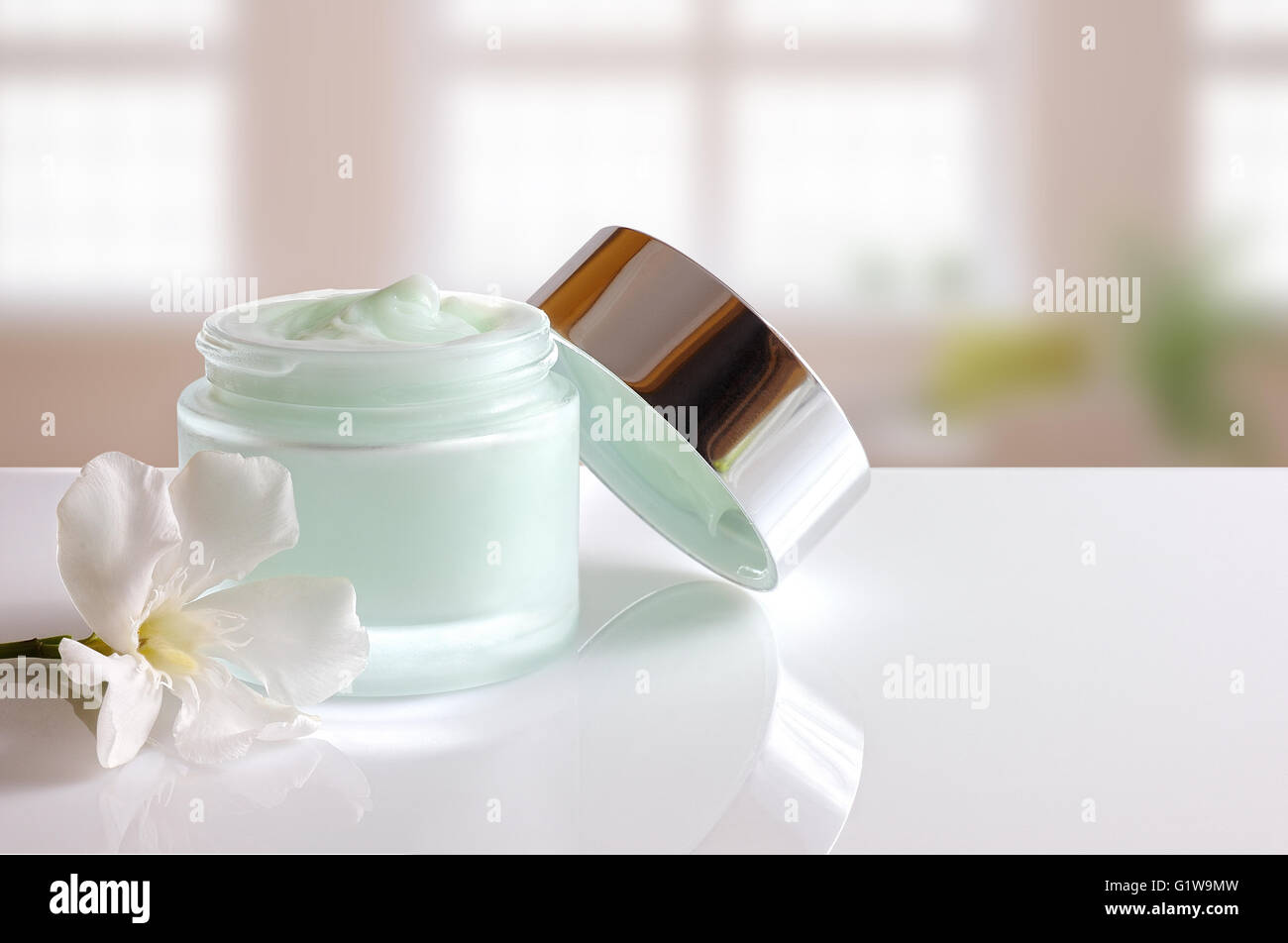 Vetro vaso aperto con trattamento viso o crema per il corpo sul tavolo bianco con coperchio e fiore. Sfondo di windows. Vista frontale. Foto Stock