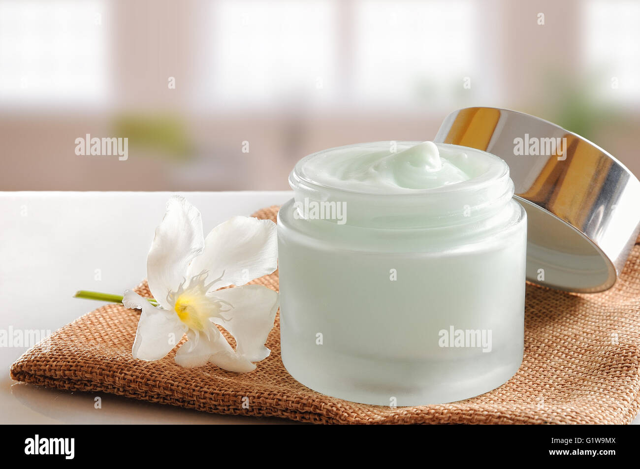 Vetro vaso aperto con viso o corpo crema sulla tela. Con coperchio e fiore.background Windows. Vista frontale. Foto Stock