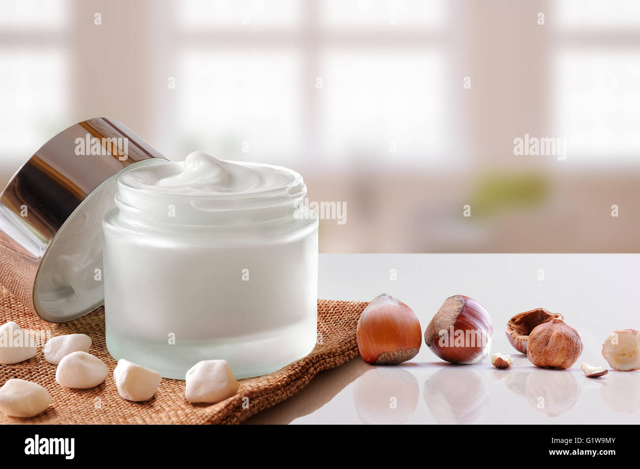 Vetro vaso aperto con viso o corpo idratante nocciole su tela. Con coperchio, con piccole pietre e nocciole.background Windows. Foto Stock