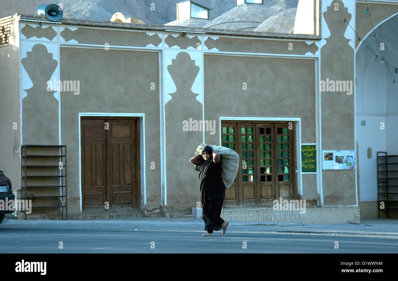 In un piccolo villaggio sul ciglio della strada una donna tradizionalmente rivestito in un chador porta un grande fascio; come altrove una donna di lavoro non viene mai fatto Foto Stock