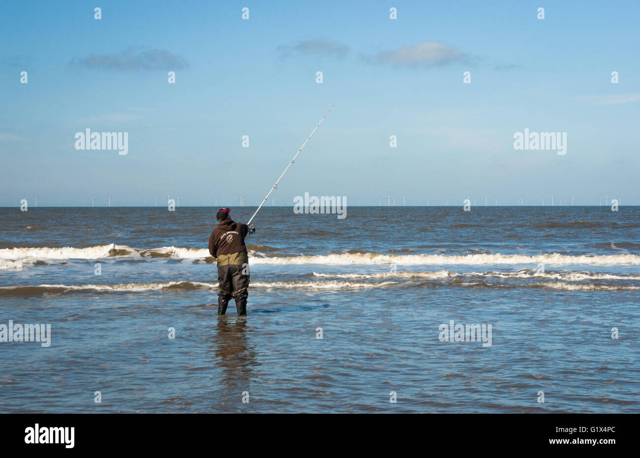 Fisher in waders in piedi knee-alta nel Mare del Nord acqua con la sua canna da pesca durante la colata della linea. Visto dal retro. Foto Stock