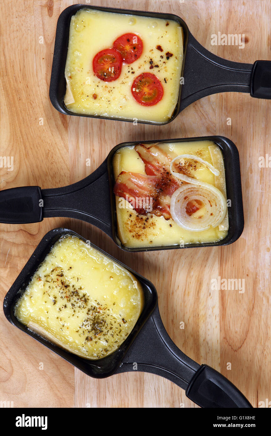 Foto di un tre vassoi Raclette con il formaggio fuso, erbe, pomodoro ciliegino, la pancetta e la cipolla. Foto Stock