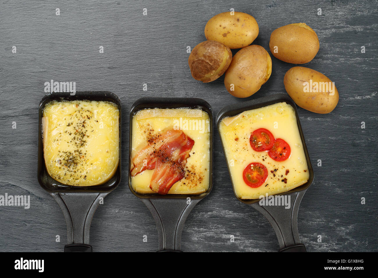 Foto di un tre vassoi Raclette con il formaggio fuso, erbe, pomodoro ciliegino, pancetta, cipolla e patate su terreni accidentati ardesia. Abbondanza di sp Foto Stock