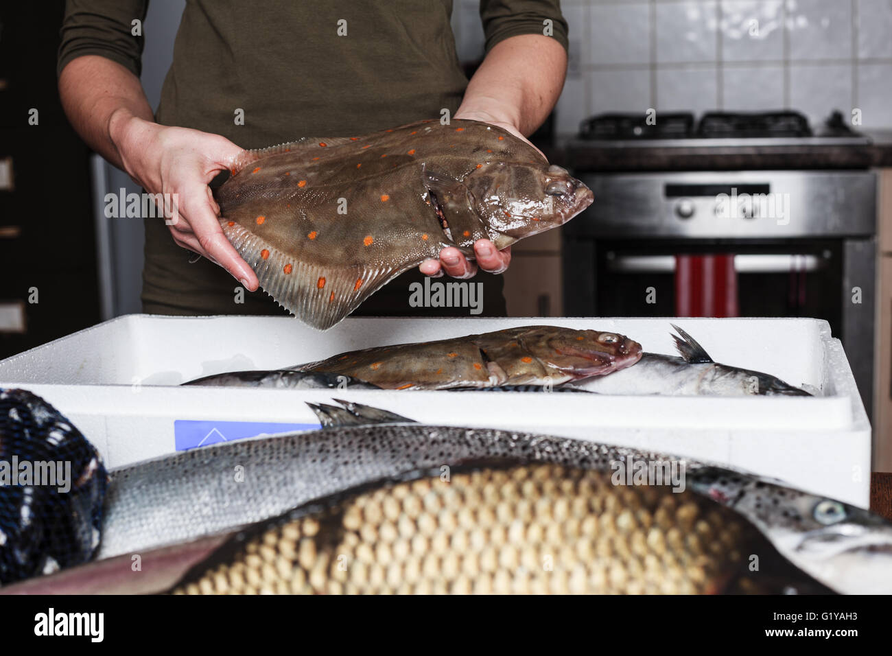Una giovane donna è nella sua cucina ed è in possesso di un passera di mare con altri tipi di pesce in una scatola di fronte a lei Foto Stock