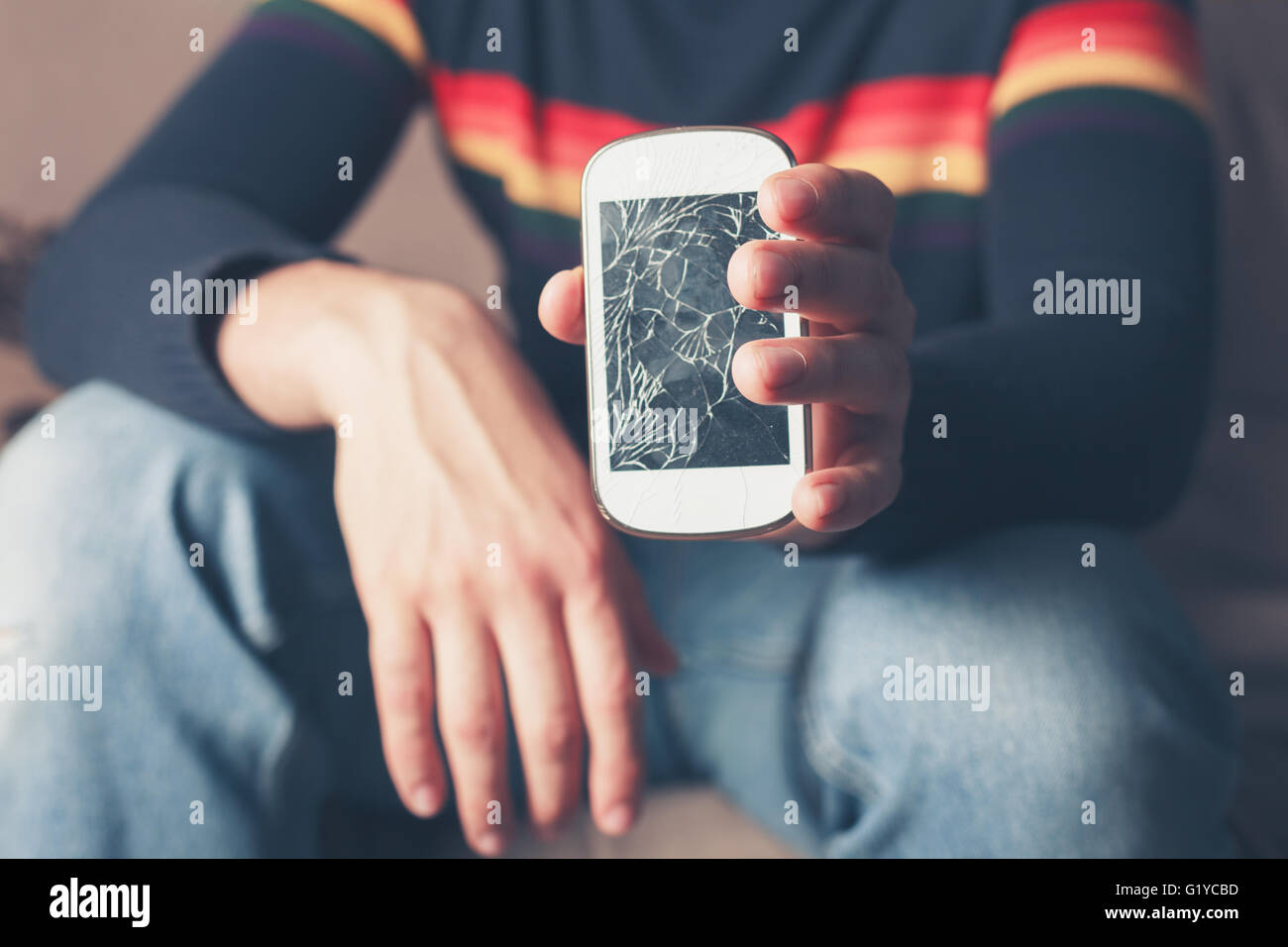 Un giovane uomo è seduto su un divano ed è in possesso di una rotta smart phone con schermo rotto Foto Stock