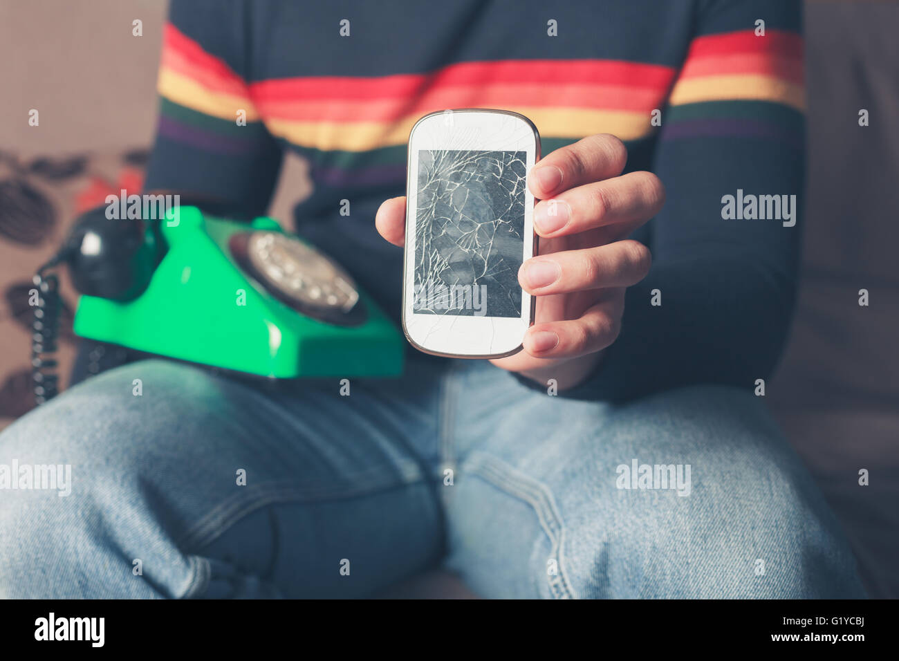 Un giovane uomo è seduto su un divano con un rotto smart phone e un vecchio telefono rotante Foto Stock