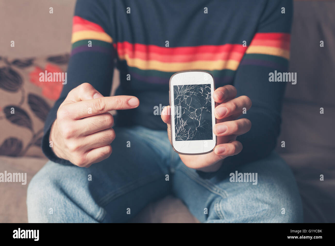 Un giovane uomo è seduto su un divano ed è rivolto a una rotta smart phone con schermo rotto Foto Stock