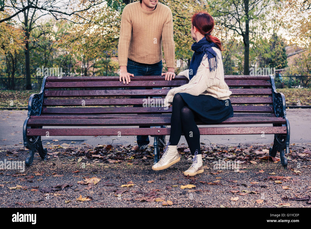 Una giovane donna è seduta su una panchina nel parco e sta parlando di un uomo che è in piedi dietro di lei Foto Stock