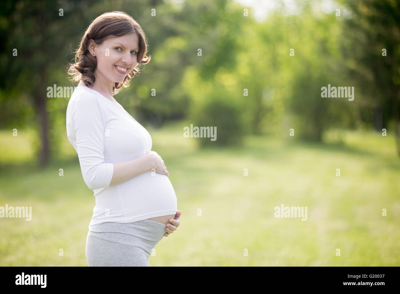 Ritratto di giovane felice modello incinta guardando la fotocamera mentre in piedi nel parco e ad accarezzarle il ventre. Futuro sorridente mom Foto Stock