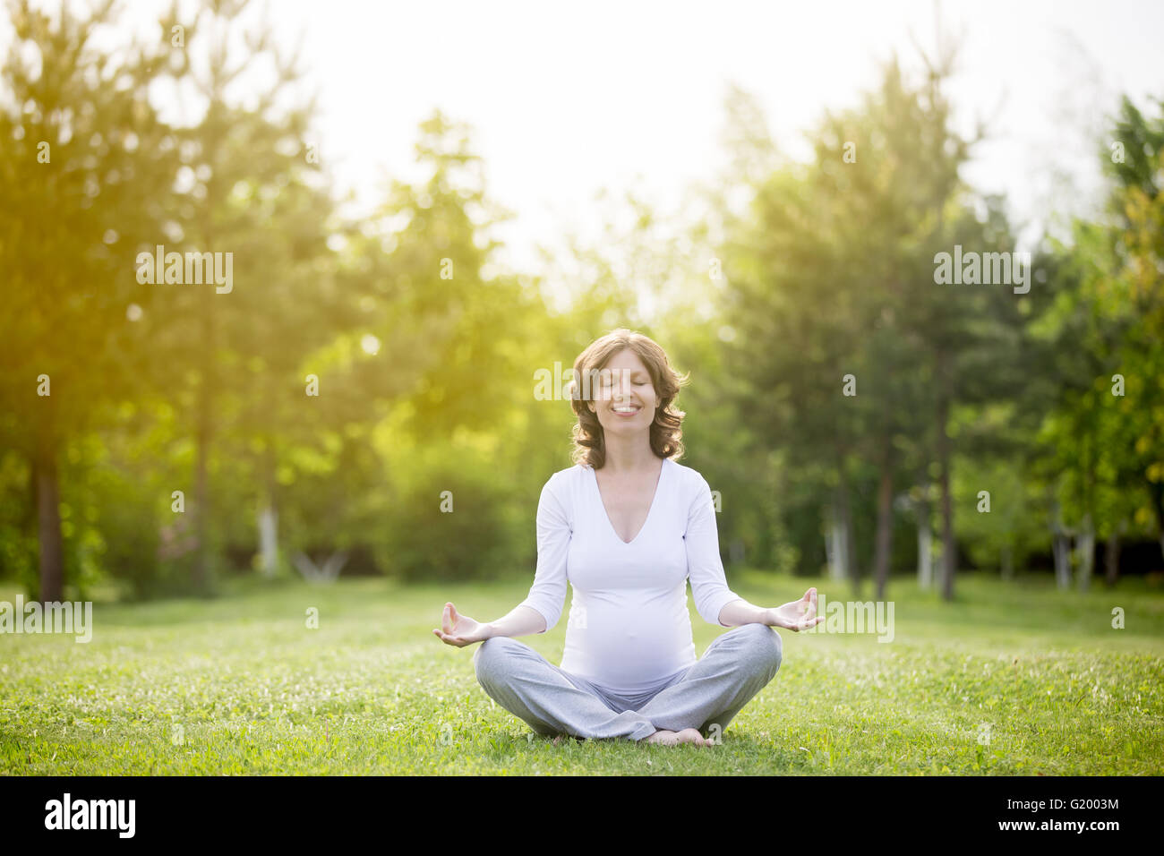 Ritratto di giovane modello incinte lavorano nel parco. Sorridente futura mamma aspetta bambino seduto a gambe incrociate e la meditazione Foto Stock