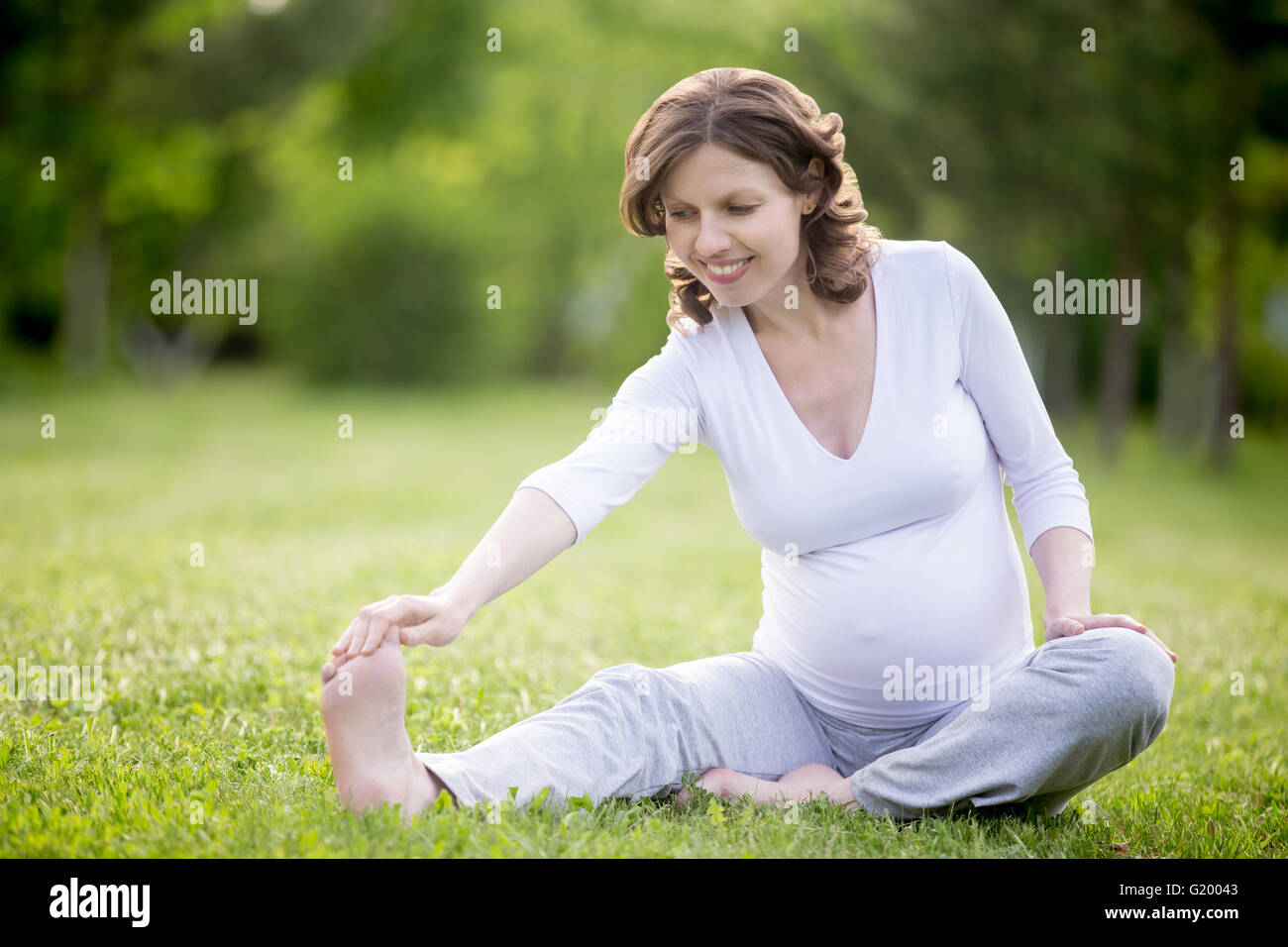 Ritratto di giovane modello incinte lavorano nel parco. Sorridente futura mamma facendo esercizi di stretching all'esterno. Seduto posteriori della coscia Foto Stock