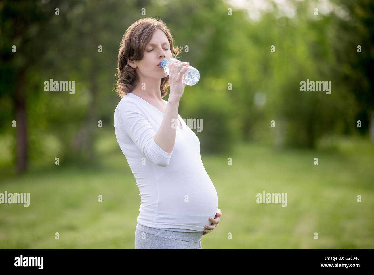 Ritratto di giovane modello gravide di bere acqua fresca con gli occhi chiusi nel parco. Futura Mamma aspetta baby tenendo la bottiglia per esterno Foto Stock