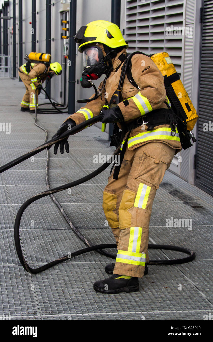 Pompiere Indossare respiratore trascina una linea hosereel su un gantry piattaforma di accesso durante una simulazione di incidente chimico in un capannone industriale. Foto Stock