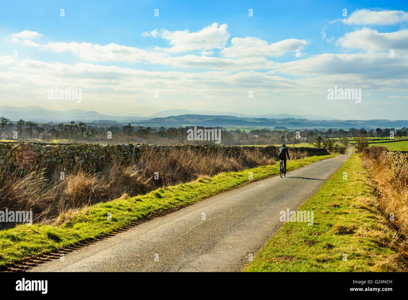 Ciclista su strada solitaria tra Kirkland e Skirwith nell'Eden Valley Cumbria con la Lakeland Fells in distanza Foto Stock