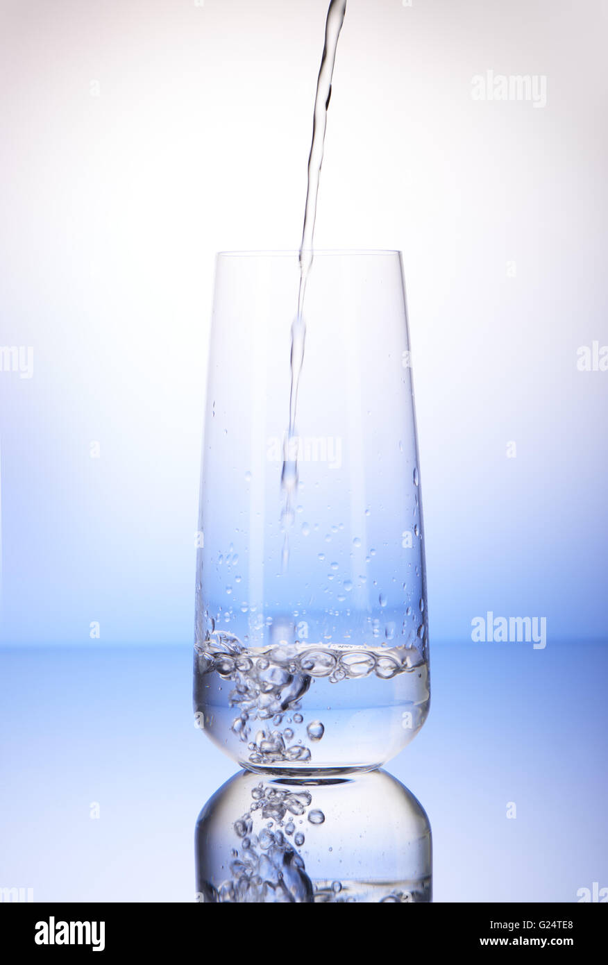 Acqua versando in uno terzi completo bicchiere con la riflessione su bianco e lo sfondo blu Foto Stock