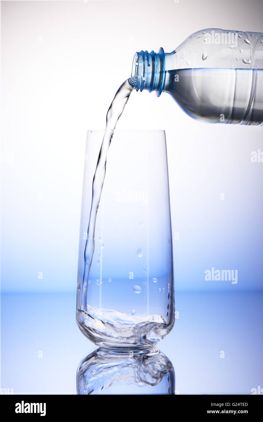 Versare acqua da bottiglie di plastica vuote nel bicchiere con la riflessione su bianco e lo sfondo blu Foto Stock