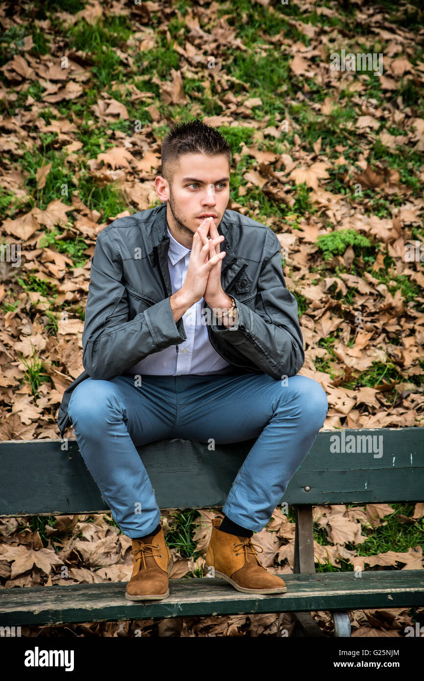 Giovane uomo bello sedersi sulla panchina nel parco in autunno, con caduta foglie tutte intorno Foto Stock